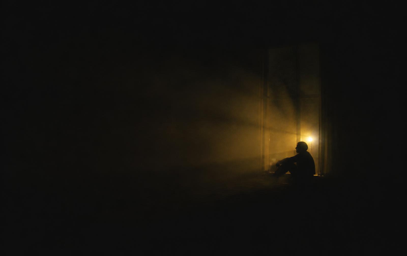 A figure sitting alone in the dark, lit by a distant amber light